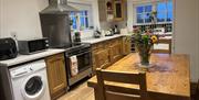 The photo is of the kitchen in Dunnaglea Cottage. There is a dining table with fresh flowers. In the background are the utilities and amenities includ