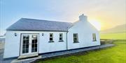 The photo is of Dunnaglea Cottage from the side. There is grass surrounding the cottage and mountains and sunrise is the background.