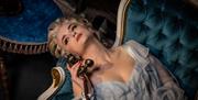 Soprano Mary McCabe reclines in a blue nightgown on a blue and white rococco chaise. She's holding an old fashioned telephone receiver and looks resig