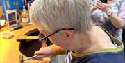 Two ladies in jewellery making workshop, one is hammering her piece of metal whilst the other takes a photograph on her phone.