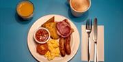 Photo of a plate with bacon, sausages, scrambled eggs, tomato, baked beans, hash brown and potato bread. Next to it is a cup of cappuccino and a glass