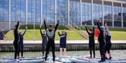 group of people dressed in wet suits balancing on paddleboards on the lake with hands in the air practicing Pilates