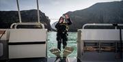 Girl on a boat in scruba diving gear falling backwards with Carrick-a-Rede Rope Bridge in the background as part of a scuba diving experience with Aqu