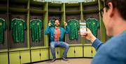 A man sitting in the player's changing rooms with football shirts beside him getting his picture taken as part of a behind the scenes guided tour of W
