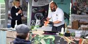 A cookery demonstration with a male chef showing salmon and a woman commentating to the crowd