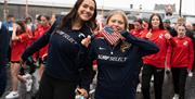 two women smile for the camera while walking in a parade - one holds a small american flag