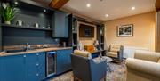 A kitchen with a sink and wine fridge extending into a lounge area with chairs, a coffee table and a TV.