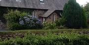 Image is of front of cottage with stone wall covered with plants and flowerbeds