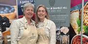 Gail from The Pantry at Bramble Hill at Balmoral Show standing in front of a display stand with a branded apron and another demonstrator