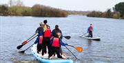 group of 6 people in wetsuits on kneeling on large SUP board on the lake with another single person paddle boarding alongside