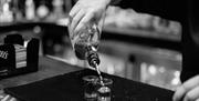 A bartender pouring liquor from a bottle into two shot glasses on a bar mat. The scene is captured in black and white, with blurred bottles and bar eq
