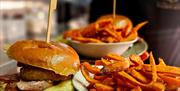 Plates of food being delivered to a table at Helena's Bistro. The dishes include gourmet burgers with crispy onion rings, served alongside sweet potat
