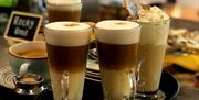 Tray of specialty coffees featuring layered lattes in tall glass mugs with frothy tops, alongside a whipped cream-topped iced coffee and a cappuccino
