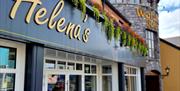 Exterior of Helena's Coffee Shop and Bistro, featuring a modern black and white facade with gold lettering. Colorful hanging plants decorate the top o