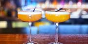 Two orange cocktails served in coupe glasses, garnished with black straws, placed on a wooden bar counter. The background is blurred, showcasing colou