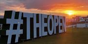 large outdoor signage at sunset in Portrush reads '#THEOPEN'