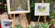 3 paintings of Northern Ireland landscape on easels, surrounded by houseplants.