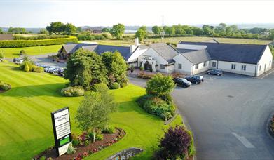 Image shows hotel and grounds with grass area and sign re hotel name. Also shows parking areas