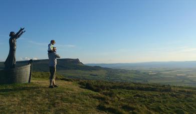 Person standing beside Manannan Mac Lir statue looking at fields and blue sky.