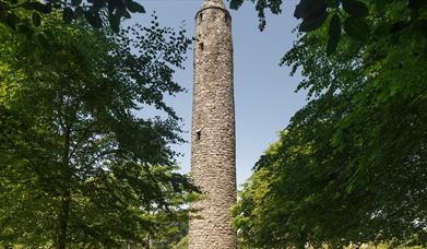 A tall thin old brick tower in a park with trees.