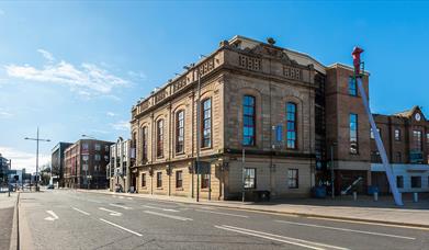 The Corn Exchange, a large light-coloured stone building with arched windows, stands on a quiet city street under a clear blue sky. There are addition
