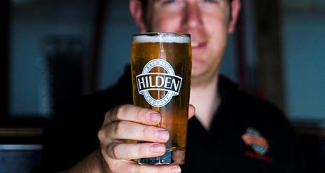 Image shows a smiling bartender holding a pint of Hilden beer