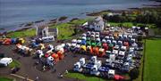 Image is an overhead aerial view of lots of lorries parked up.  Several of them are brightly coloured and you can see the sea and the coastline in the
