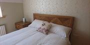 A double bed, made with white bedding and a decorative cushion. Next to the bed is a wooden bedside table. On the table is a bedside light in a silver