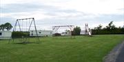 Image is of swings, climbing frame and a slide in a field near to caravans
