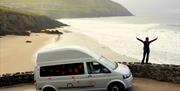 Image is of campervan parked with a lady standing on stone wall with arms outstretched. View of a beach and the sea