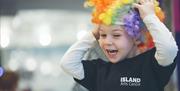 Image shows a laughing child wearing a brightly coloured hair attachment and wearing an Island Arts Centre t-shirt