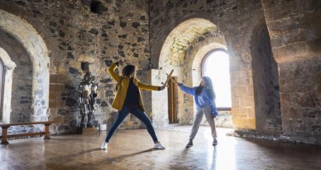 A woman and child having a pretend sword fight in the room at the top of the castle keep with the sun beaming through the window behind.