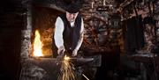 Photo of a gent dressed in old fashioned clothing in his workshop. There is a flaming fire and sparks are flying as he works on a piece of iron