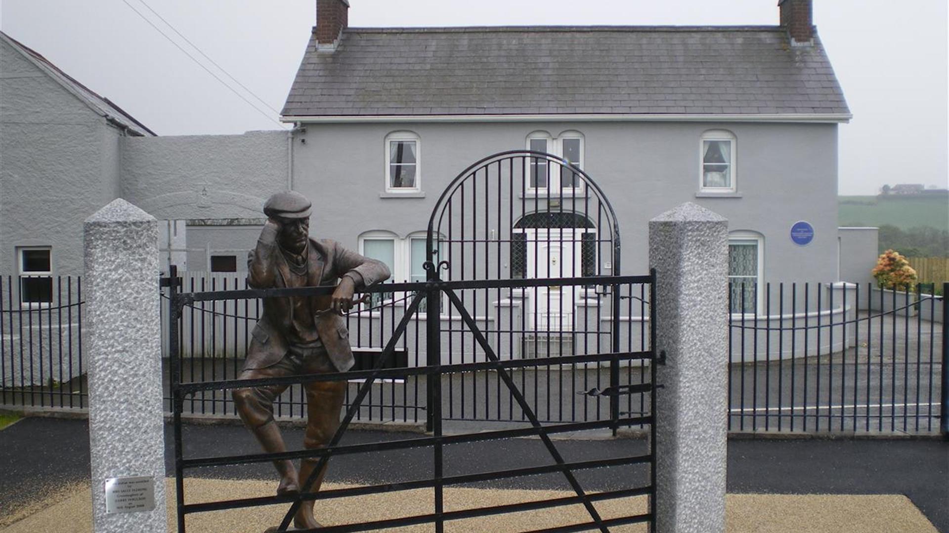Image is of Harry Ferguson bronze statue leaning on gate  in front of his family home