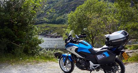 Image shows motorcycle parked at scenic area with view of river between trees