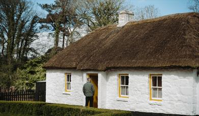 A person standing at the entrance of a white stone cottage with yellow rimmed windows and a thatch roof