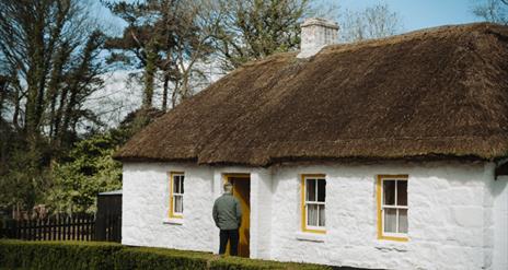 A person standing at the entrance of a white stone cottage with yellow rimmed windows and a thatch roof