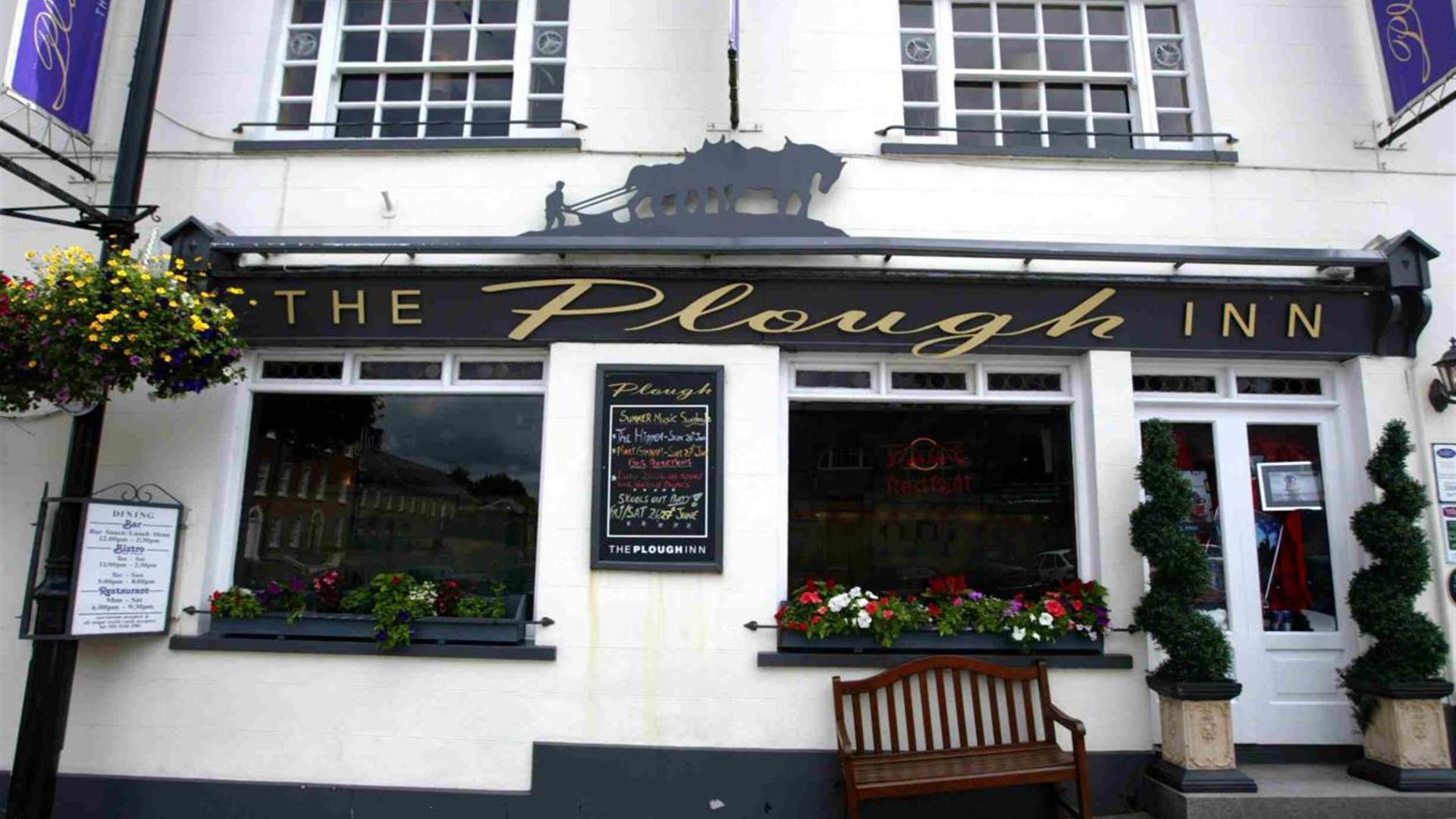 Image is of the outside of the restaurant with a wooden bench outside one of the windows and bay trees at either side of the front door