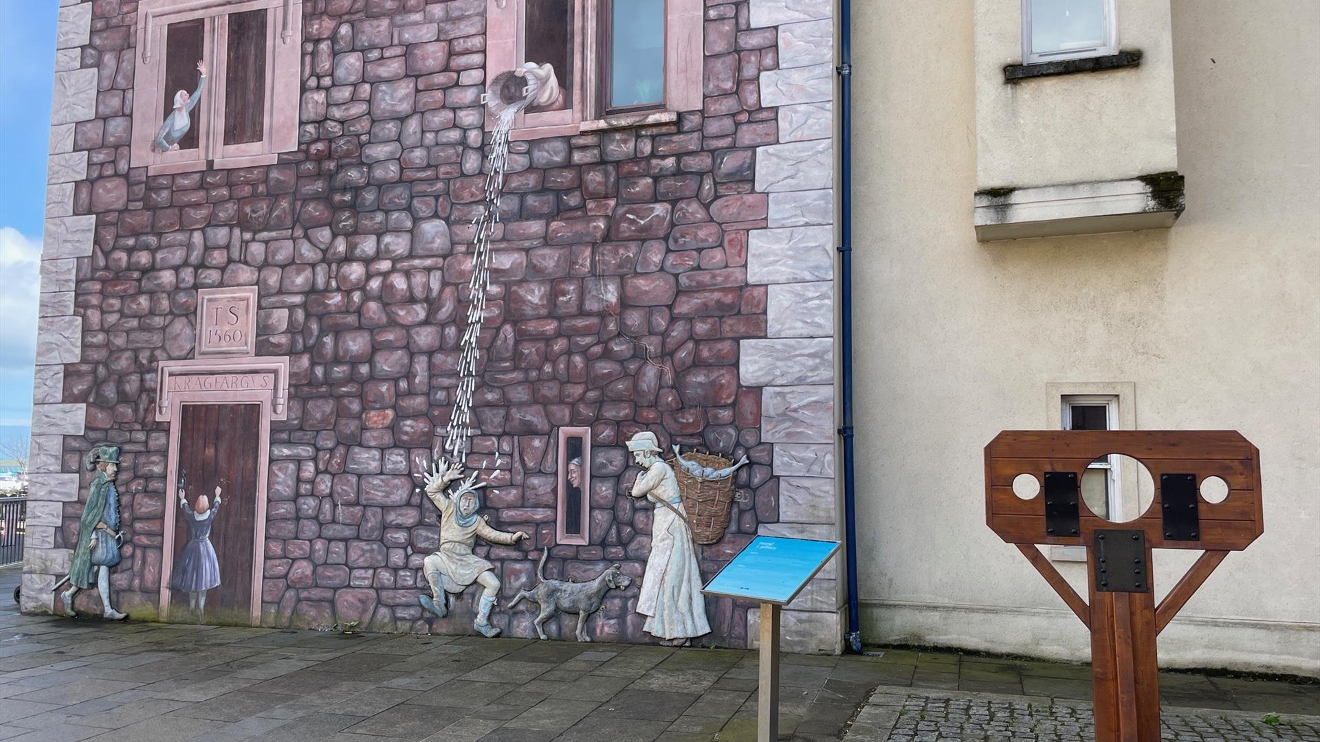 Medieval wall mural in Carrickfergus, with stocks beside it.