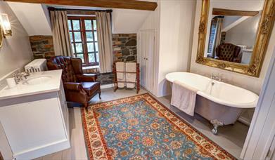Image shows large bathroom with roll top bath and large mirror above it. Large rug. armchair, wash basin with storage underneath
