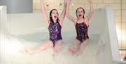 Image shows 2 girls with their hands in the air and going down a slide in the swimming pool