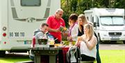 Image shows family and friends sitting at picnic table with caravan in the background