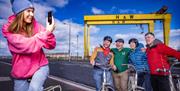 A young woman in a pink hoodie snaps a photograph of Steven from Hometown Tours with three guests posing on their bikes under the shipyard cranes.