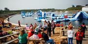 Image shows lake with inflatables and area around it with picnic tables and people standing around watching the activities