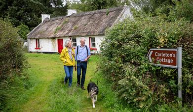 A mature couple and their dog walk through grass away from an Irish thatched cottage.