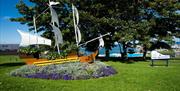 A photo of a boat sculpture set within a garden display located on grassland beside the seafront area