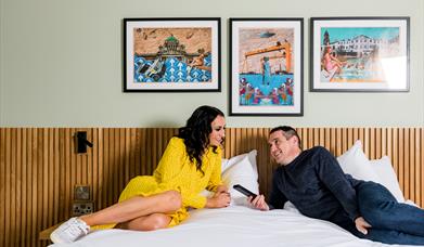 Image shows a man and woman lounging on a double bed with paintings of Northern Ireland on the background wall