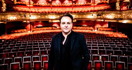 Opera Director Cameron Menzies standing on stage at the Grand Opera House with the red velvet seating behind him
