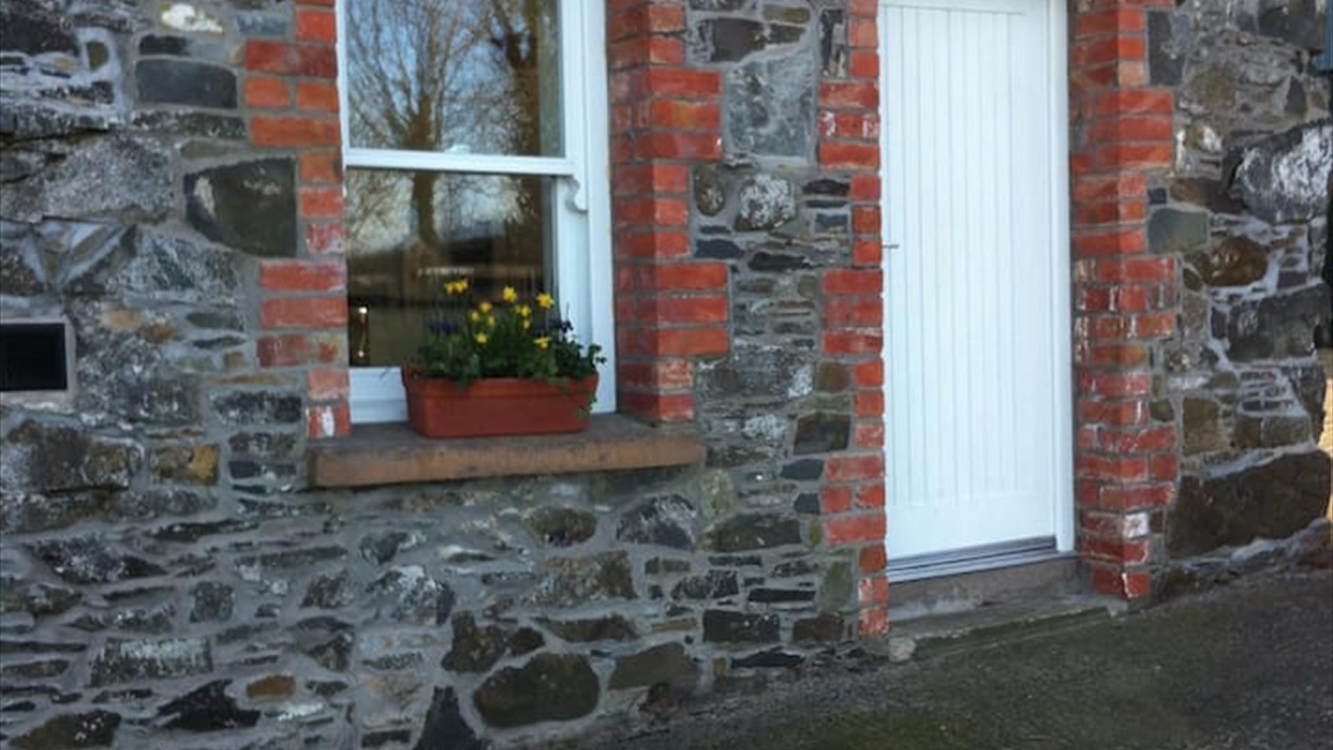 Image is of a stone cottage with a white front door and flowerbed on the window sill