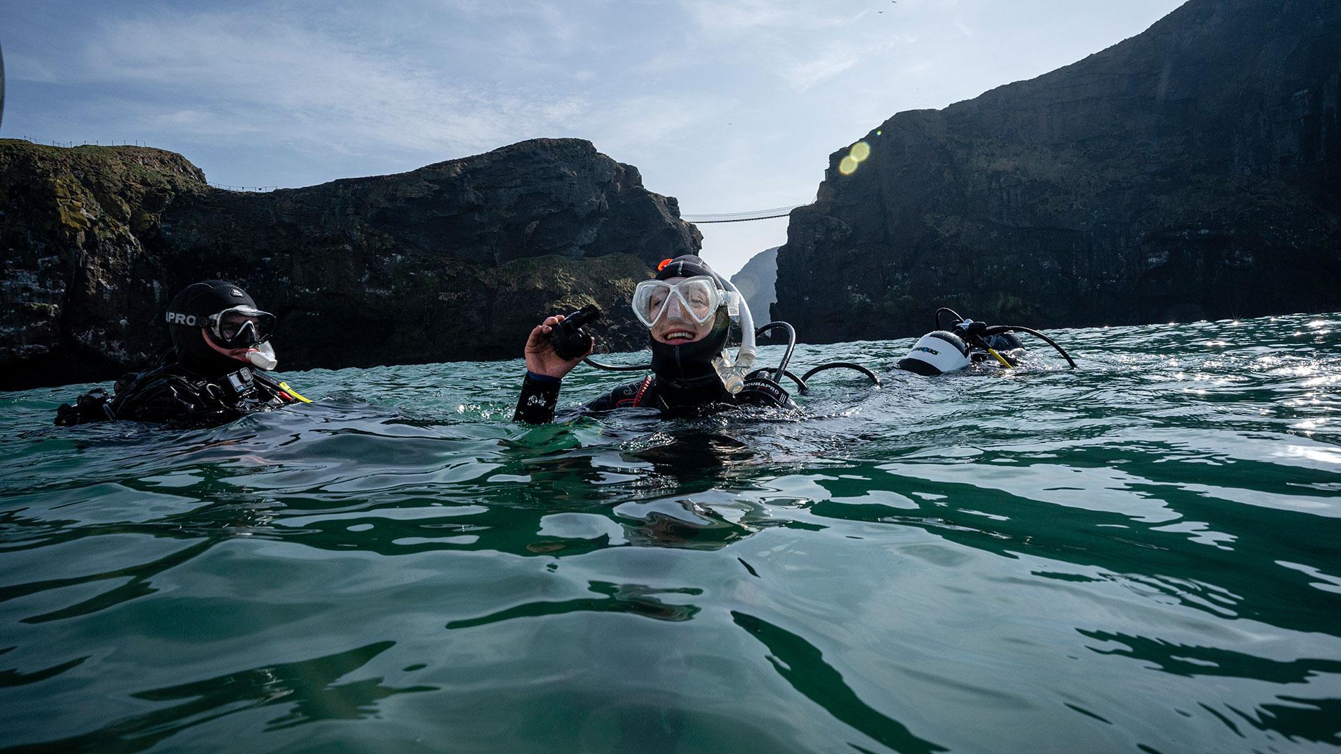 Group in wetsuits in the water with Carrick-a-Rede Rope Bridge in the background as part of a scuba diving experience with Aquaholics